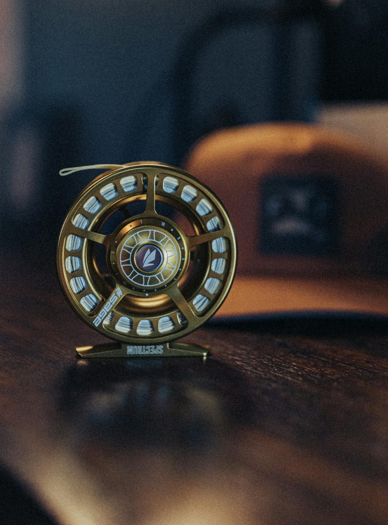 Home Detailed shot of a gold fly fishing reel placed on a wooden surface with a blurred cap in the background.